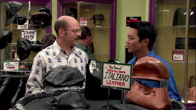 A bald mustachioed man in a patterned shirt speaks with a younger man at a hat and leather goods store counter, with a sign prominently advertising '100% Full Grain Italiano Leather.' The scene appears to be from a TV sitcom set in a retail shop.
