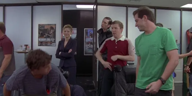 A group of people gathered in an office or workplace setting, with a woman standing with arms crossed looking skeptical while several men appear to be reacting to something. A GamePro magazine cover featuring Resident Evil 4 is visible on the wall in the background.