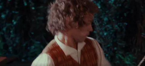 A curly-haired hobbit character in a rustic vest looks downward with a curious or cautious expression, set against a lush green outdoor background typical of the Shire. The scene appears to be from The Lord of the Rings or The Hobbit film series.