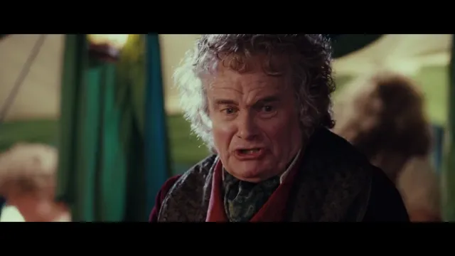 An elderly hobbit character with curly gray hair and a red scarf appears agitated or confrontational, captured in a tense close-up scene from a fantasy film. This is Bilbo Baggins from The Lord of the Rings, likely during the birthday party scene.